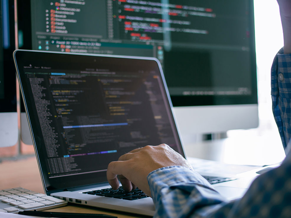 A programmer typing code one a laptop computer with a larger display in the background